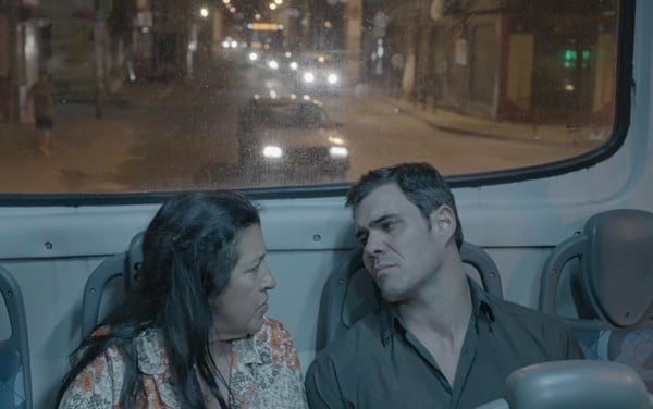 Na imagem da novela Amor de Mãe, Regina Casé (Lurdes) de camisa florida e Juliano Cazarré (Magno) com camisa social escura estão sentados em um ônibus à noite no Rio de Janeiro