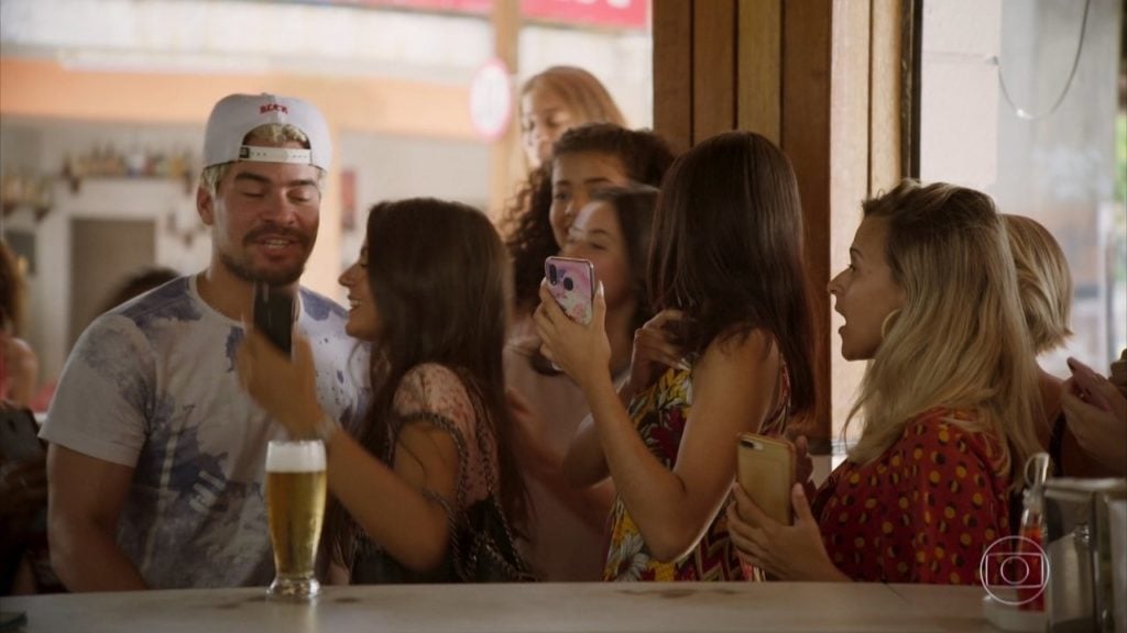 Em cena da novela Amor de Mãe, o ator Thiago Martins como o personagem Ryan está em um bar sendo abordado por algumas garotas com celular na mão