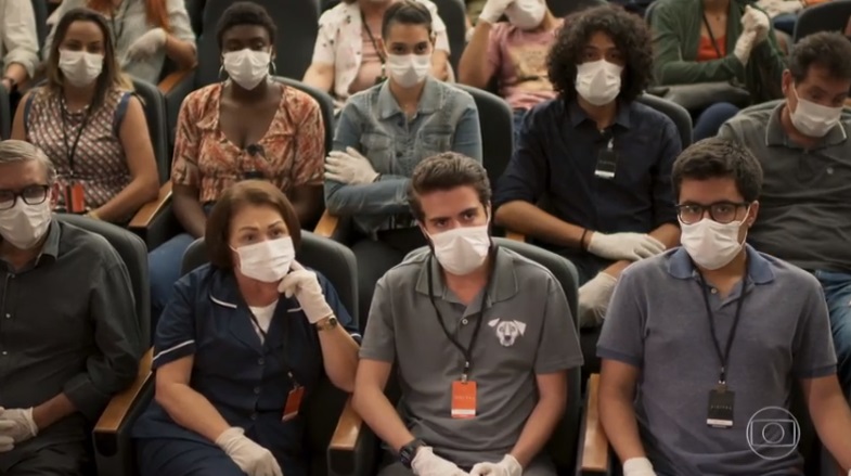 Pessoas diversas estão sentadas em um mini teatro em cena de Salve-se Quem Puder usando máscaras nos rostos e luvas