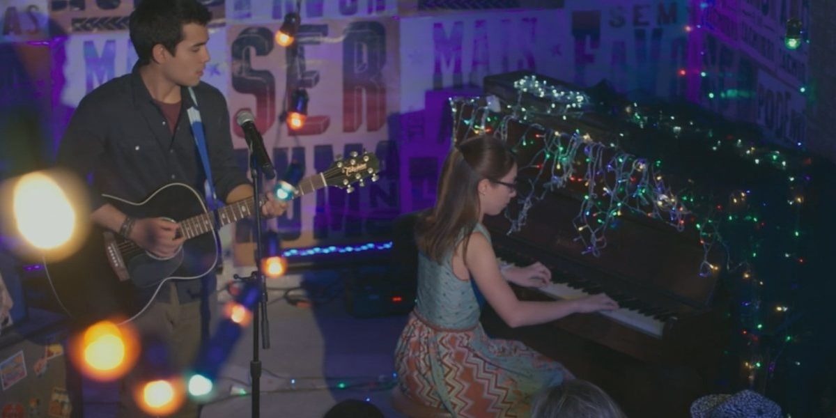 Na foto da cena da novela Malhação - Viva a Diferença em ambiente escuro iluminado por diversas luzes de natal, Benê esta no piano e Guto em pé cantando e tocando em frente a plateia