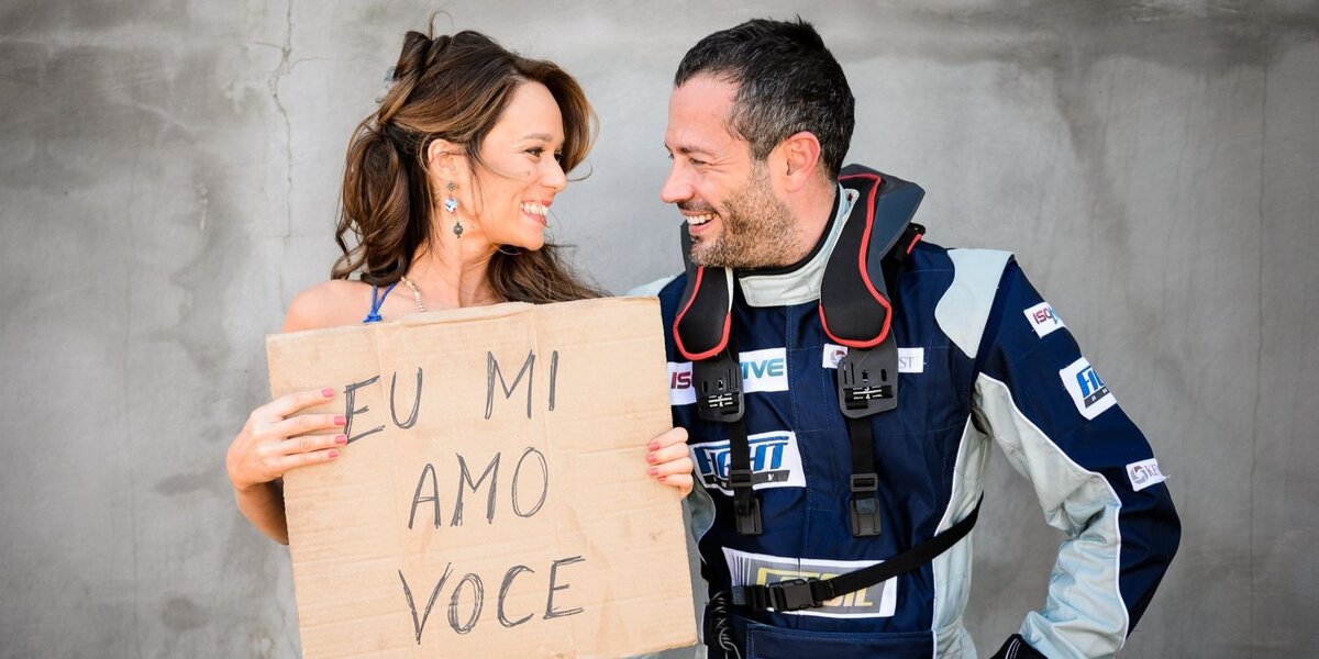 Tancinha e Apolo, vestido de piloto de corrida, se olham sorrindo e ela carrega cartaz escrito "eu mi amo você" em bastidor da novela Haja Coração