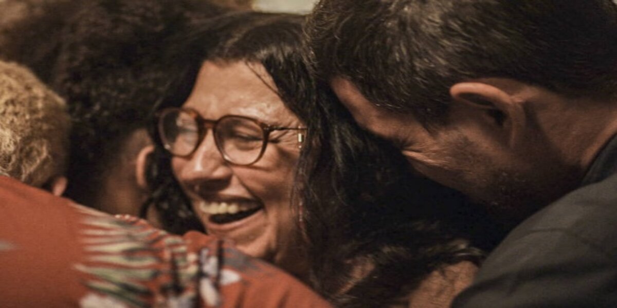 Lurdes sendo abraçada enquanto sorri usando óculos na novela Amor de Mãe