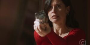 Regina usa vestido vermelho e segura uma arma em cena da novela Cara e Coragem