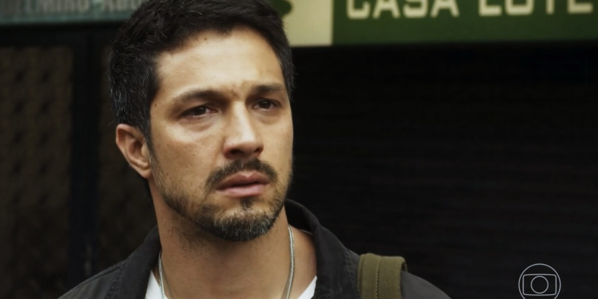 Oto em cena da novela Travessia com olhar triste, usa camisa azul social, camisa branca, seu tom de pele é branco tem barba pequena e cabeço preto curto