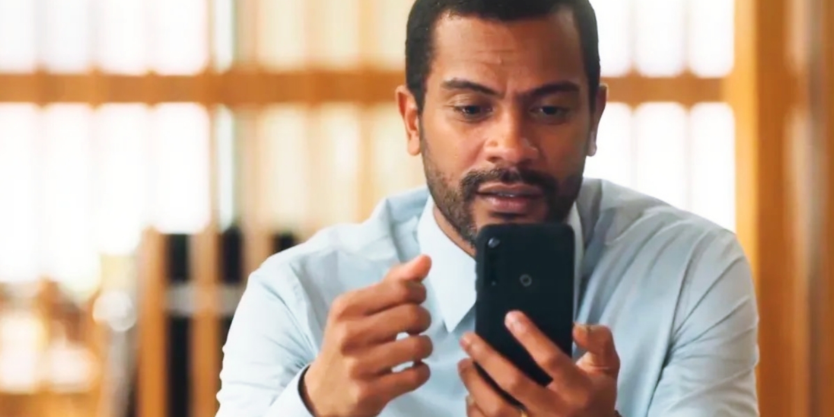 Ben, homem negro de cabelo preto bem curto, usando camiseta social azul clara, segura o celular preto com expressão de surpresa na novela Vai na Fé