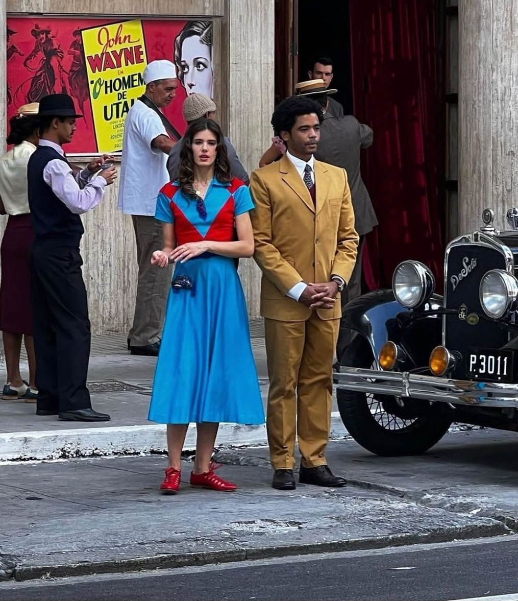 Atores Camila Queiroz e Diogo Almeida estão no rua perto de um carro preto antigo ela usa vestido longo azul com vermelho e ele está vestido com terno e calça amarelo escuro