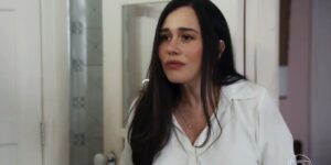 Guida é uma mulher branca, que usa cabelo longo liso preto e uma camiseta social branca e está em cena da novela Travessia