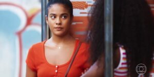 Jeniffer da novela Vai na Fé é uma mulher jovem, negra que está usando uma camiseta laranja e o cabelo está preso