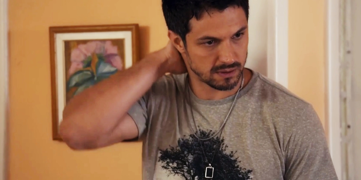 Oto usando camiseta cinza com estampa de uma árvore coloca a mão na nuca em preocupação na novela Travessia