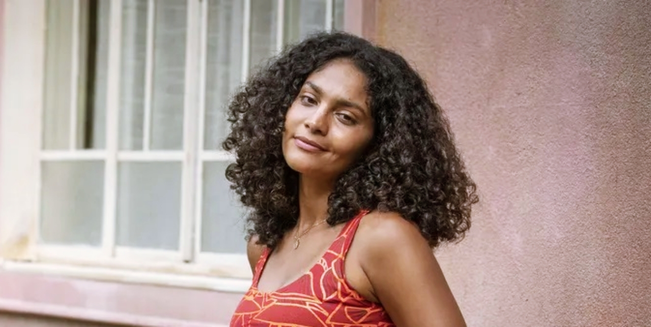 Bárbara Reis vestindo um vestido vermelho com listras brancas, com cabelo curto cacheado castanho escuro em bastidor de Terra e Paixão