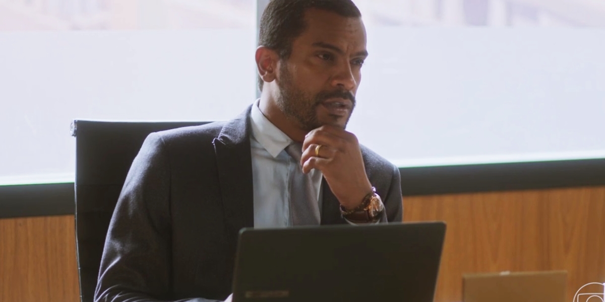 Ben em cena da novela Vai na Fé está sentado frente a uma mesa de escritório, tem em frente um notebook aberto e usa terno cinza, camisa branca