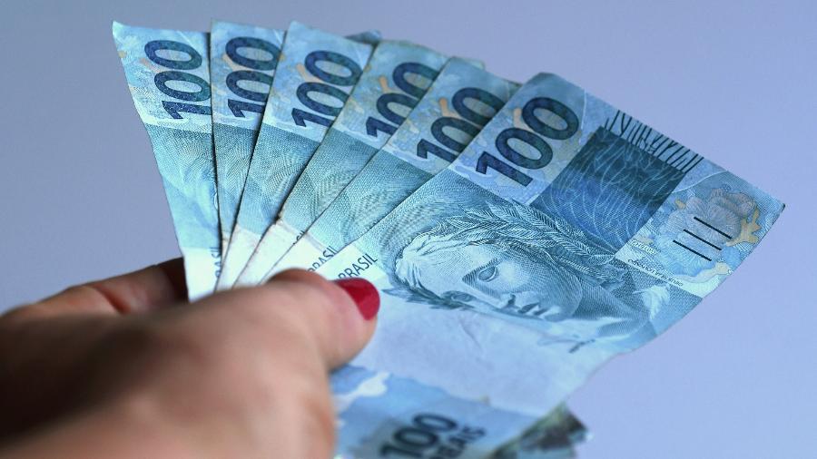 Notas de 100 reais seguradas por uma mão (Foto: Reprodução, Uol) 